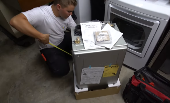 A person kneels on a concrete floor, measuring a water heater unit next to a washing machine and red tool bag, with installation manuals and parts on top of the unit.