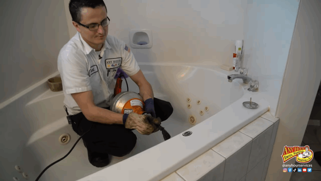 A plumber in uniform uses a drain cleaning machine on a bathtub drain in a bathroom with white tiles and a whirlpool tub.