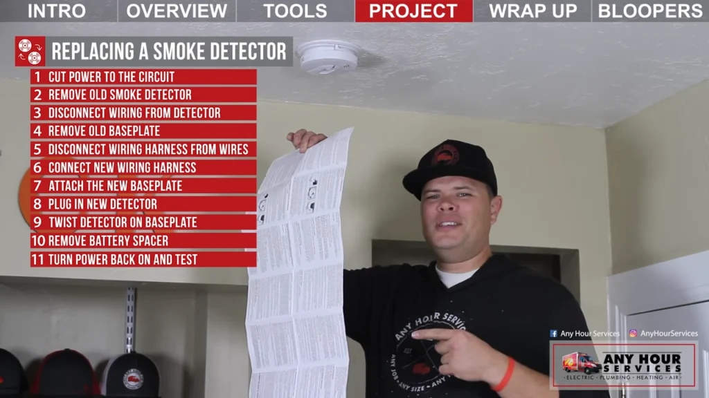 %Focuskw% | Any Hour Services A man points at a long instruction sheet under a ceiling-mounted smoke detector, with numbered replacement steps listed on a screen overlay.