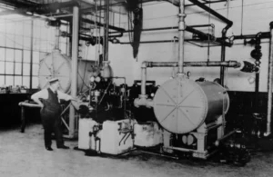A man stands next to large industrial machinery with pipes and cylindrical tanks in a factory setting, likely from the early to mid-20th century.