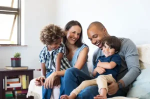 A family of four sits on a couch, smiling and laughing together in a brightly lit living room, enjoying quality time that highlights the value adds of togetherness.