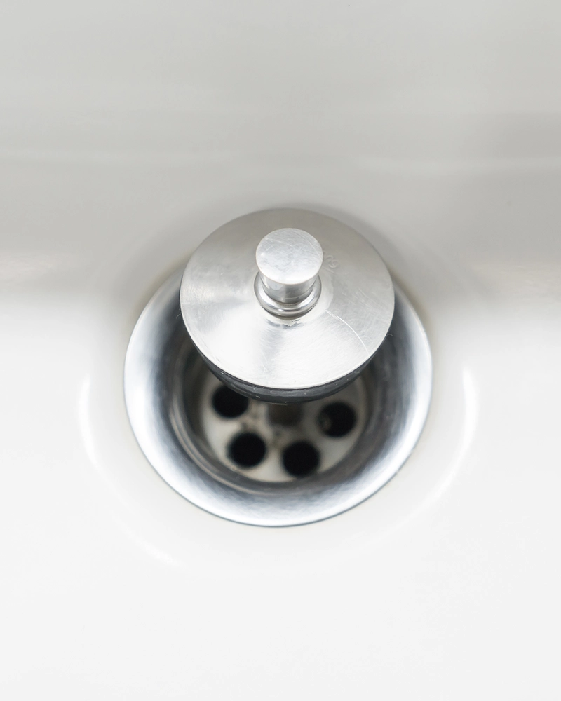 Close-up of a chrome sink drain with a shiny metal stopper in a white basin. The clean, minimalist scene conveys a sense of simplicity and modern hygiene.