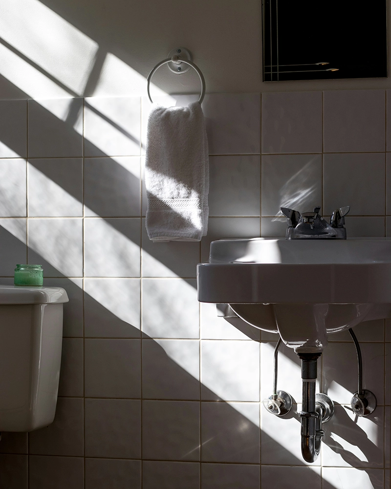 Sunlight streams through a window, casting shadows across a tiled bathroom wall. A white towel hangs on a ring beside a sink, creating a serene, calm mood.