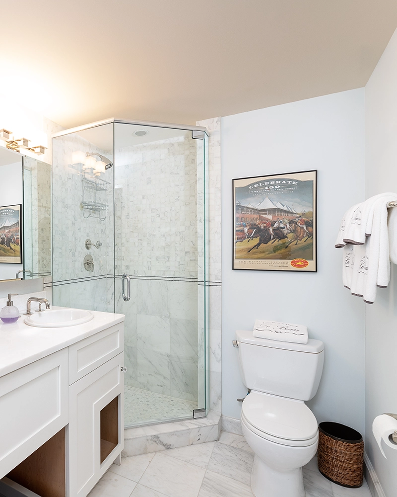 A clean, modern bathroom with a glass-enclosed shower, marble tiles, white vanity, toilet, and bathroom lighting.