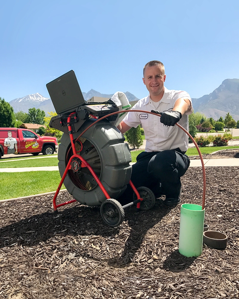 A worker in a white uniform kneels beside sewer inspection equipment on a landscaped lawn. A red service van is in the background, with mountains under a clear blue sky.