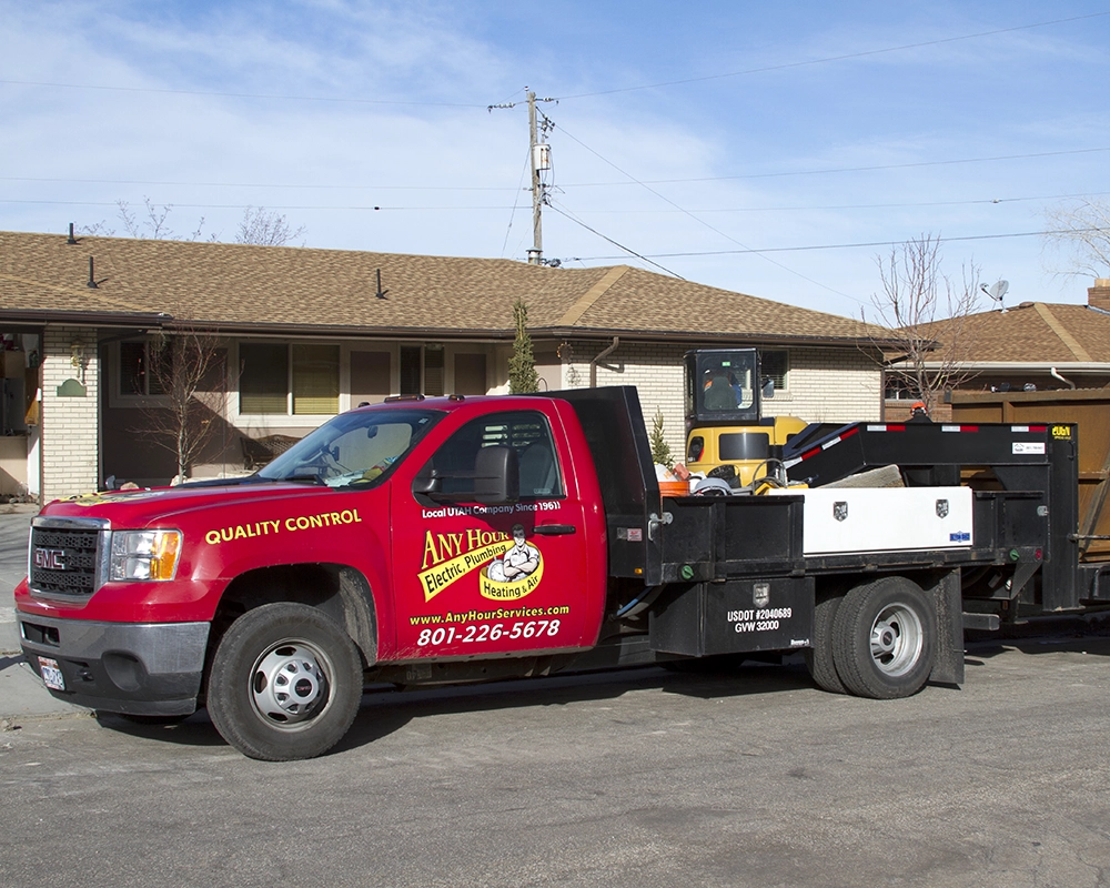 A red truck with Any Hour Electric, Plumbing, Heating & Air logo and contact info parked in front of a house on a sunny day. Trailer attached.