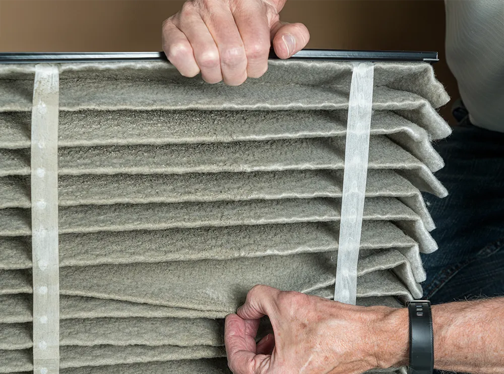 Close-up of hands holding a dirty air filter with visible dust in pleats. The person's hands suggest maintenance. The tone is practical and focused.