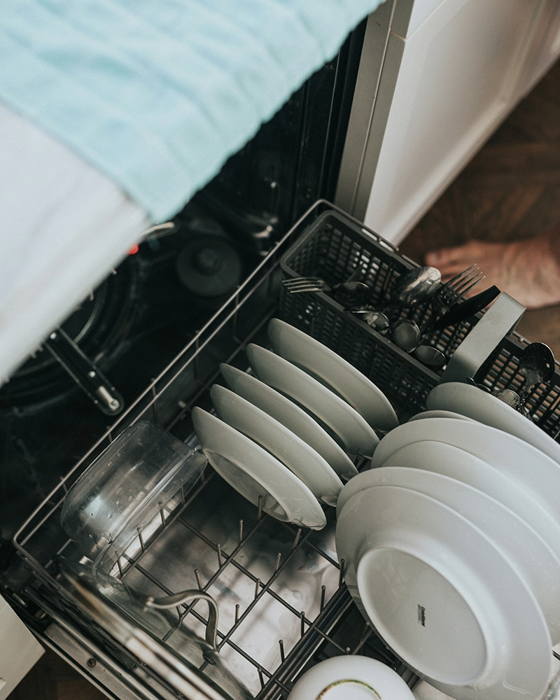 Open dishwasher with neatly stacked white plates, bowls, and utensils inside. The setting conveys a sense of cleanliness and order.