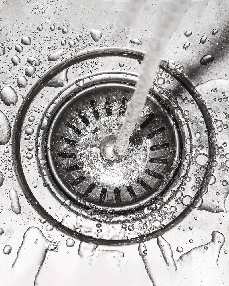 Close-up of a stainless steel sink with water flowing into the drain, splashing and creating droplets.