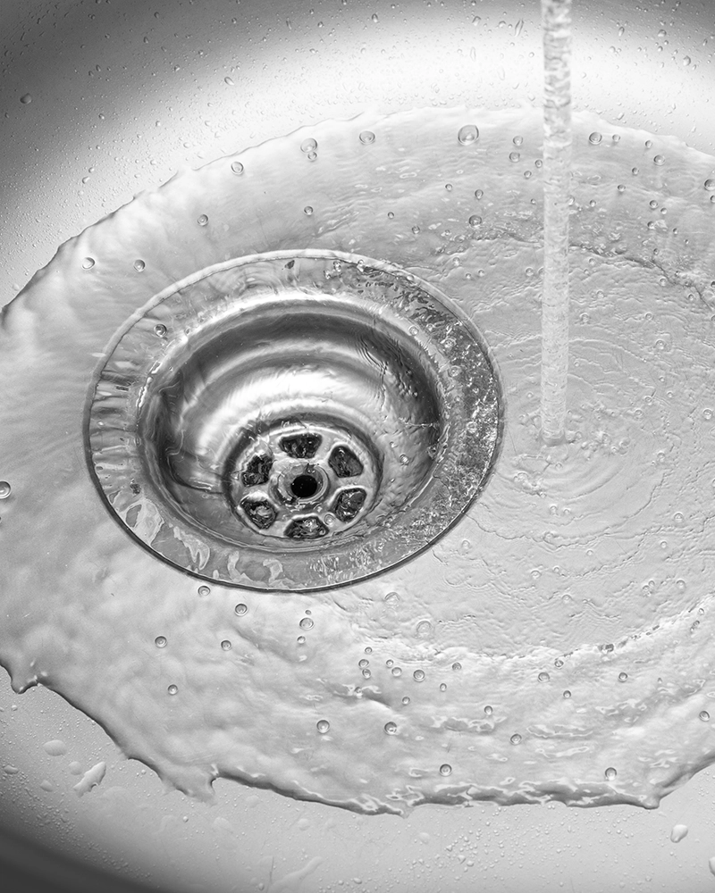 A close-up of a stainless steel sink with water flowing into the drain. The water creates swirling patterns, conveying a sense of motion and cleanliness.