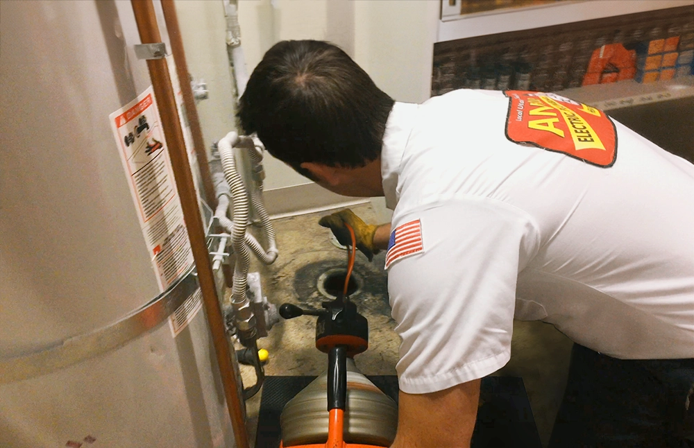 A technician in a white shirt with a logo kneels beside a water heater, using tools to work on drains/sewers. The setting is industrial and focused.