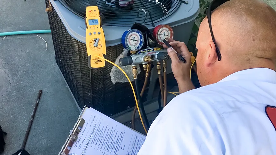 A technician inspects HVAC gauges and digital meter on an air conditioning unit, holding a clipboard with documents, focused and working diligently.