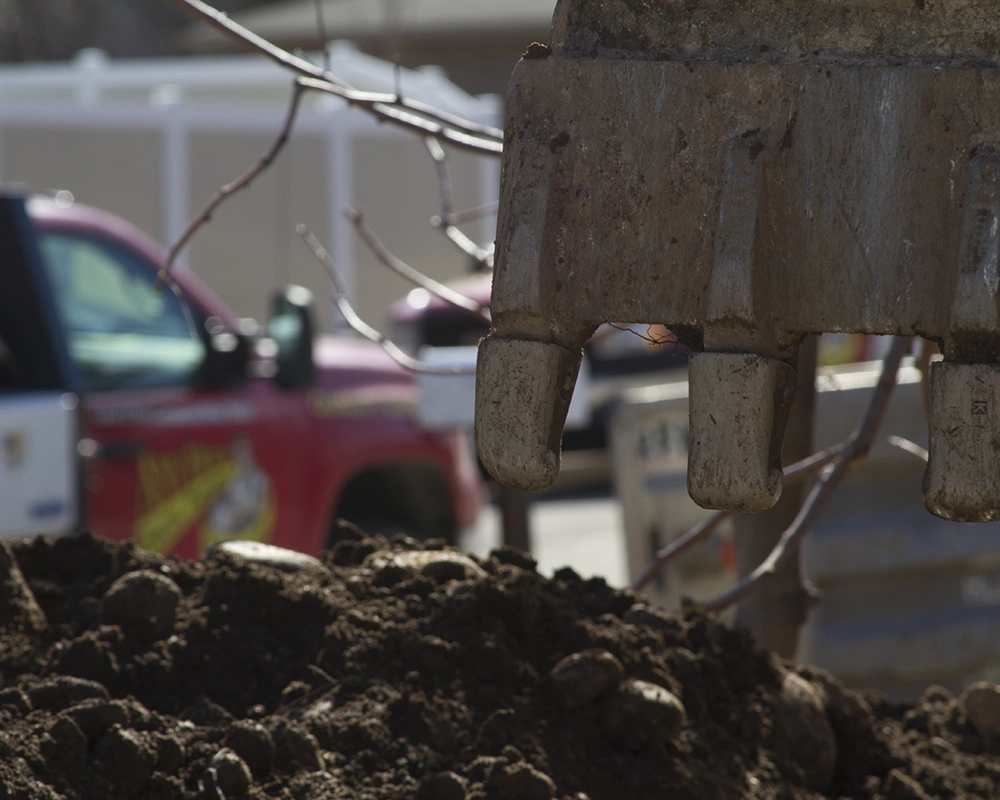 Close-up of a construction site, showing an excavator bucket above a pile of dirt. A red utility truck is blurred in the background, conveying a busy work scene.