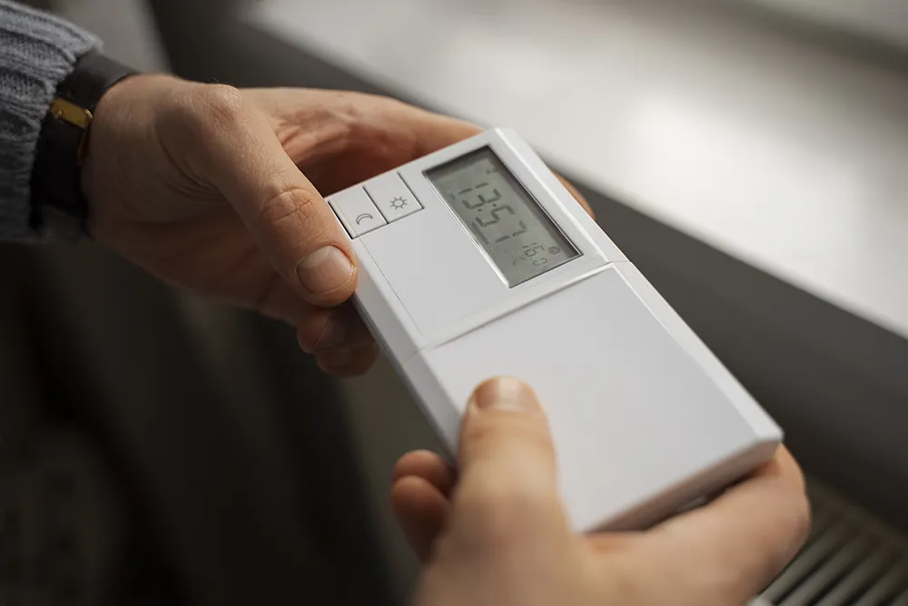 Close-up of hands adjusting a digital thermostat, displaying the temperature. The setting is indoors with soft lighting, conveying a calm, focused tone.