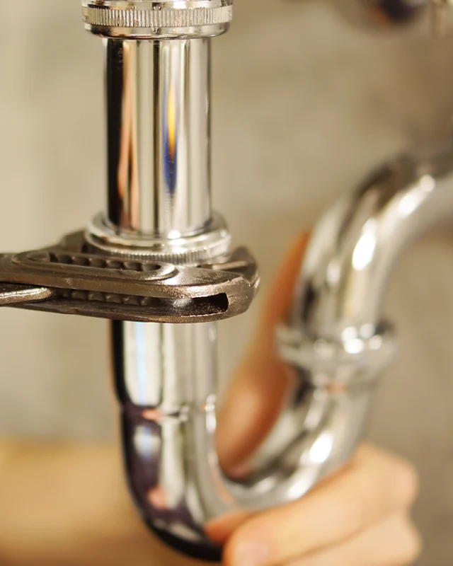 Close-up of a hand tightening a shiny chrome pipe with a wrench, against a blurred background. The image conveys a sense of focus and precision.