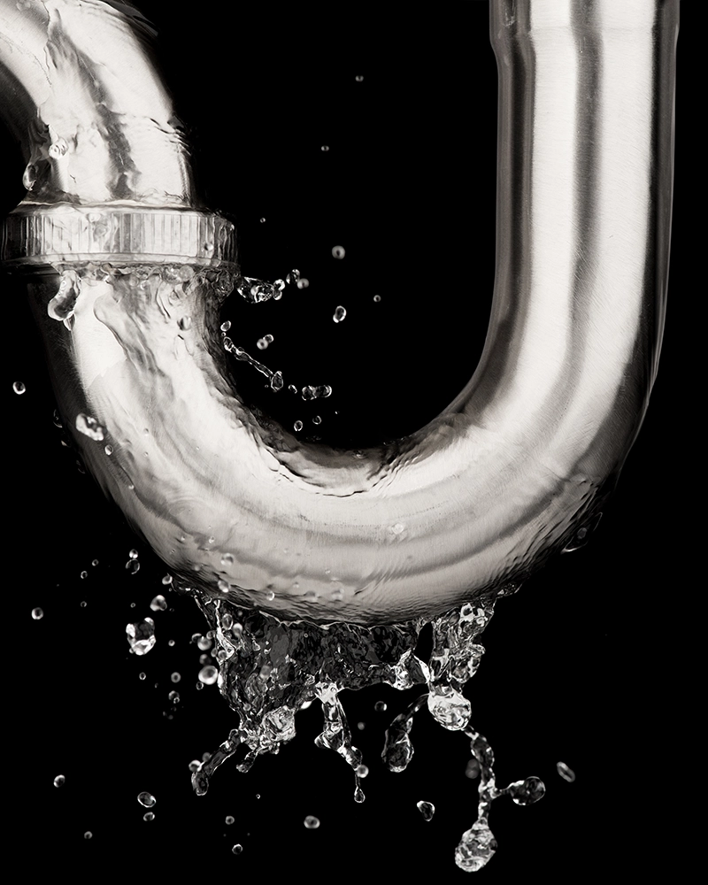 A shiny metal pipe with a U-bend leaks water, splashing droplets against a black background, conveying urgency and a need for repair.