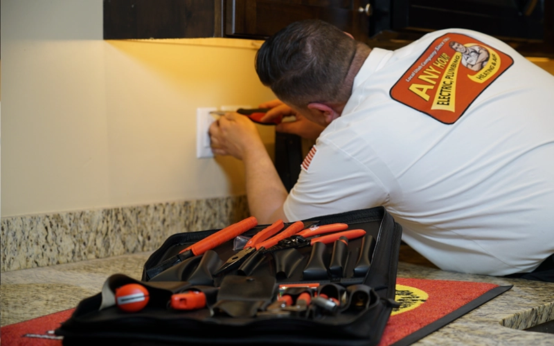 Electrician in a uniform with company logo fixes a wall outlet on a granite countertop, with a spread of red-handled tools in the foreground.