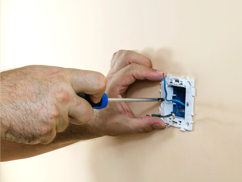 Hands using a screwdriver to install or repair an electrical outlet on a beige wall. The blue wires and open wall socket indicate home maintenance work.