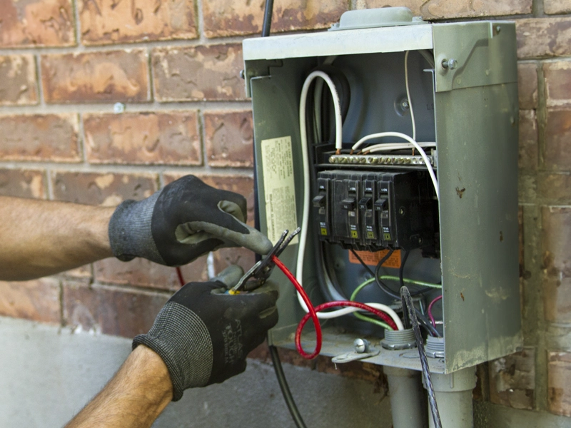 Electrician's gloved hands using pliers to work on a circuit breaker box mounted on a brick wall. Open panel reveals wires and breakers. Focused tone.