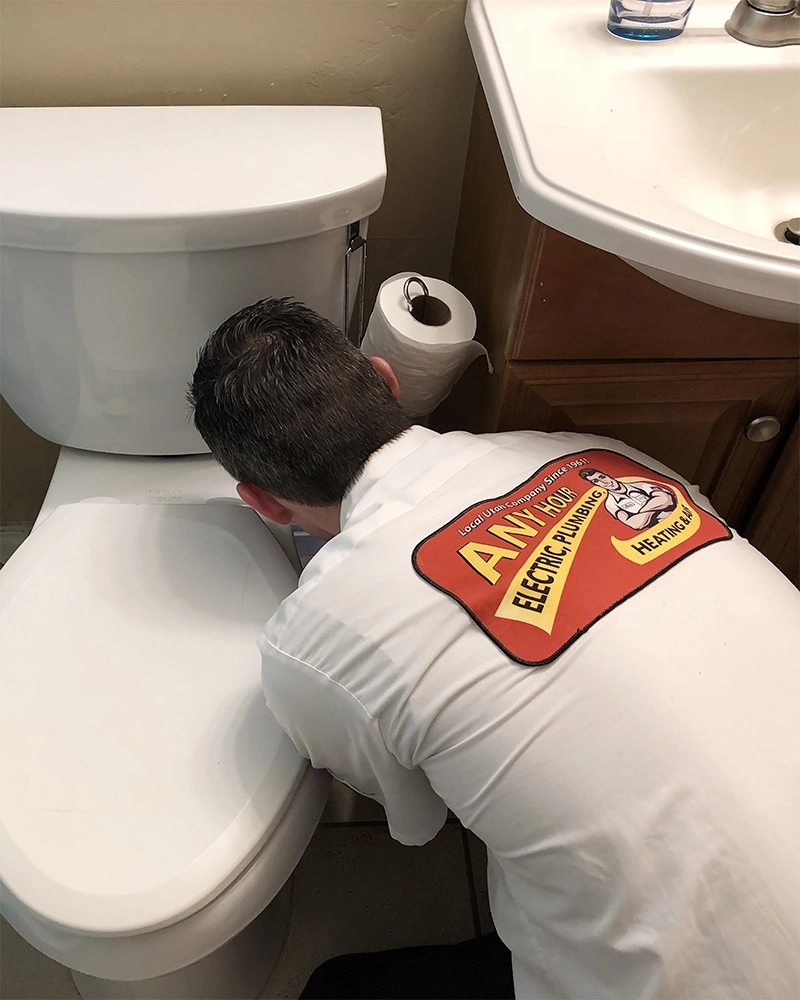 A plumber in a white uniform with a logo is inspecting a toilet. The setting is a bathroom with a sink and toilet paper roll nearby. The mood is professional.