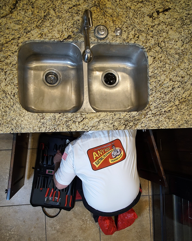 A dual kitchen sink with granite countertop is shown. A plumber in a branded shirt and red shoe covers works under the sink, with tools spread on the floor.