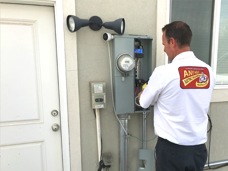 Electrician in a branded shirt works on an open electrical panel outside a building. The scene conveys professionalism and focus.