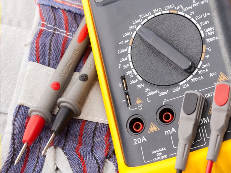 Close-up of a digital multimeter with red and gray test probes. The device rests on a colorful, textured fabric, highlighting precision and utility.