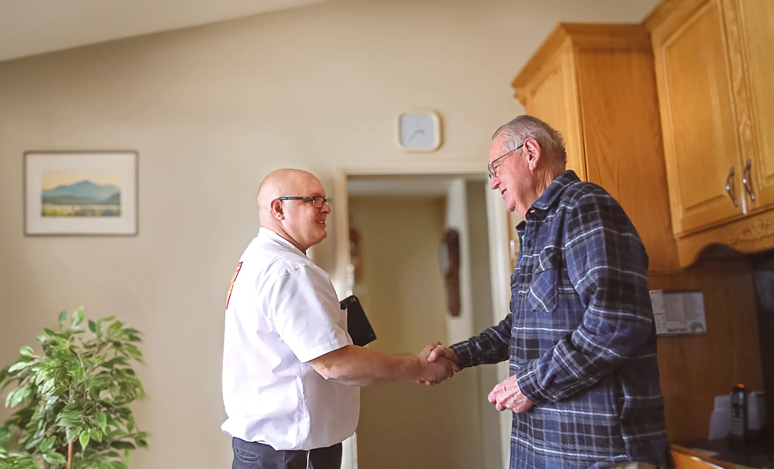 An elderly man and technician smiling and shaking hands in a cozy kitchen. One man wears a plaid shirt, the other a white shirt. Warm, friendly atmosphere.