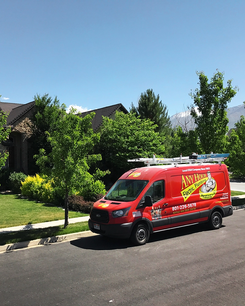 Red utility van with Any Hour Electric Plumbing logo parked on a suburban street. Lush green trees and a house with a stone facade are in the background.