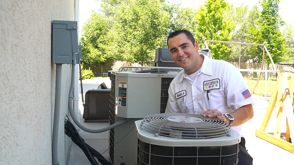 Smiling technician in a white uniform inspects outdoor air conditioning units on a sunny day. Lush trees and a yard are visible in the background.