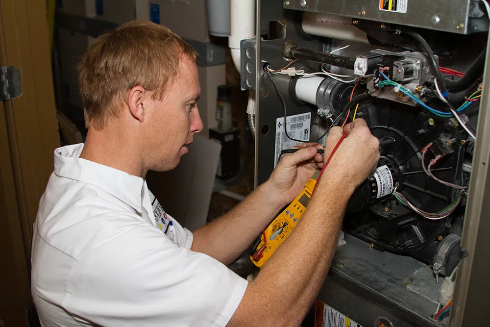 A technician in a white shirt uses a multimeter to check wires inside a furnace. Focused expression, conveying precision and technical skill in a workshop.