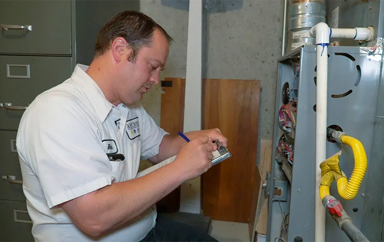 A technician in a white shirt takes notes on a clipboard while inspecting a furnace in a utility room, displaying focus and professionalism.