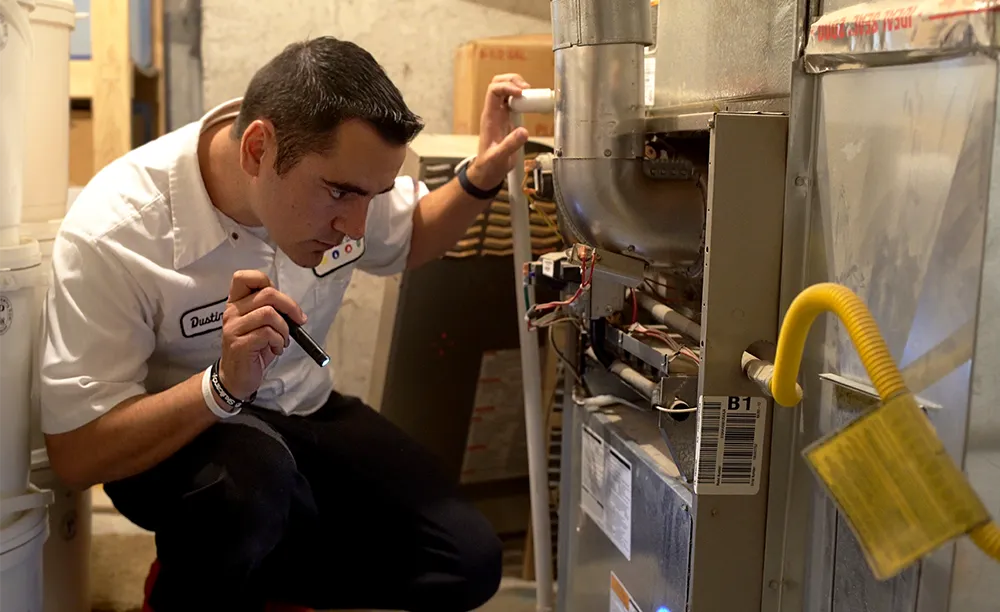 A technician in a white uniform inspects a furnace with a flashlight in a dimly lit room. He looks focused and attentive. Heating maintenance.