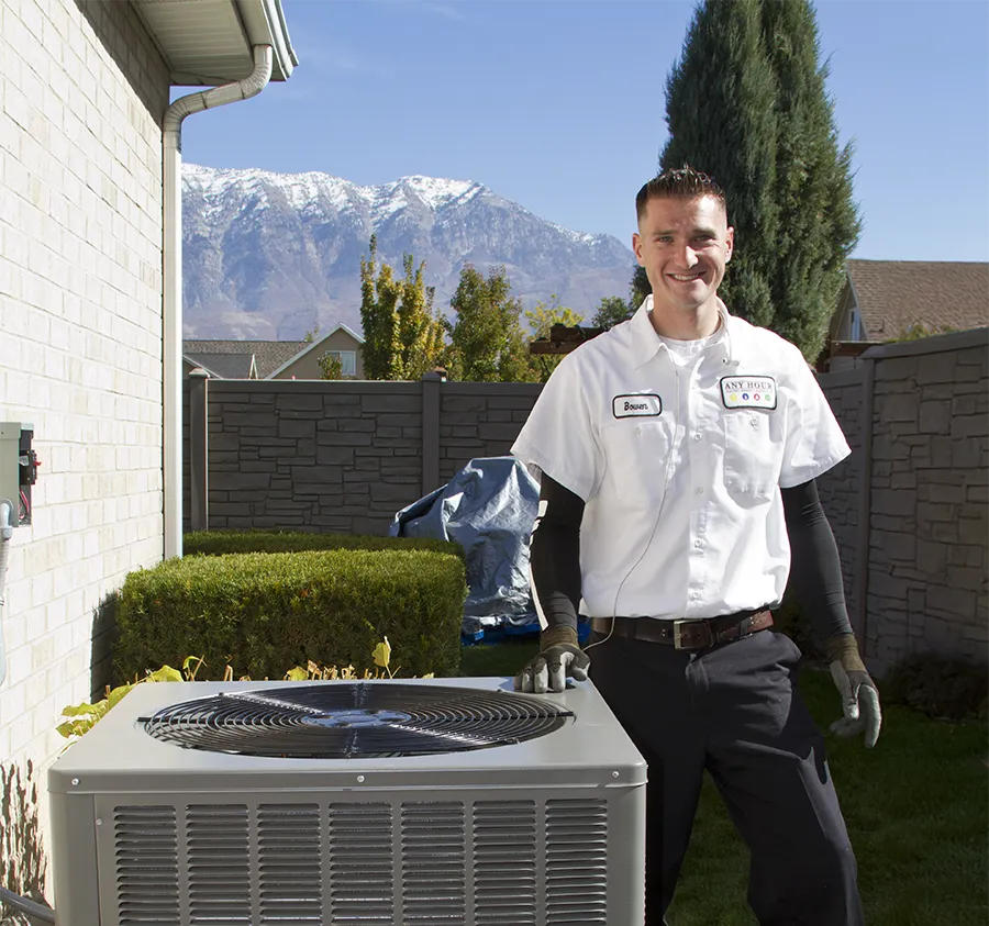 A technician in a white uniform stands outside next to an air conditioning unit, smiling. Snow-capped mountains and trees are visible in the background, suggesting a clear, sunny day.