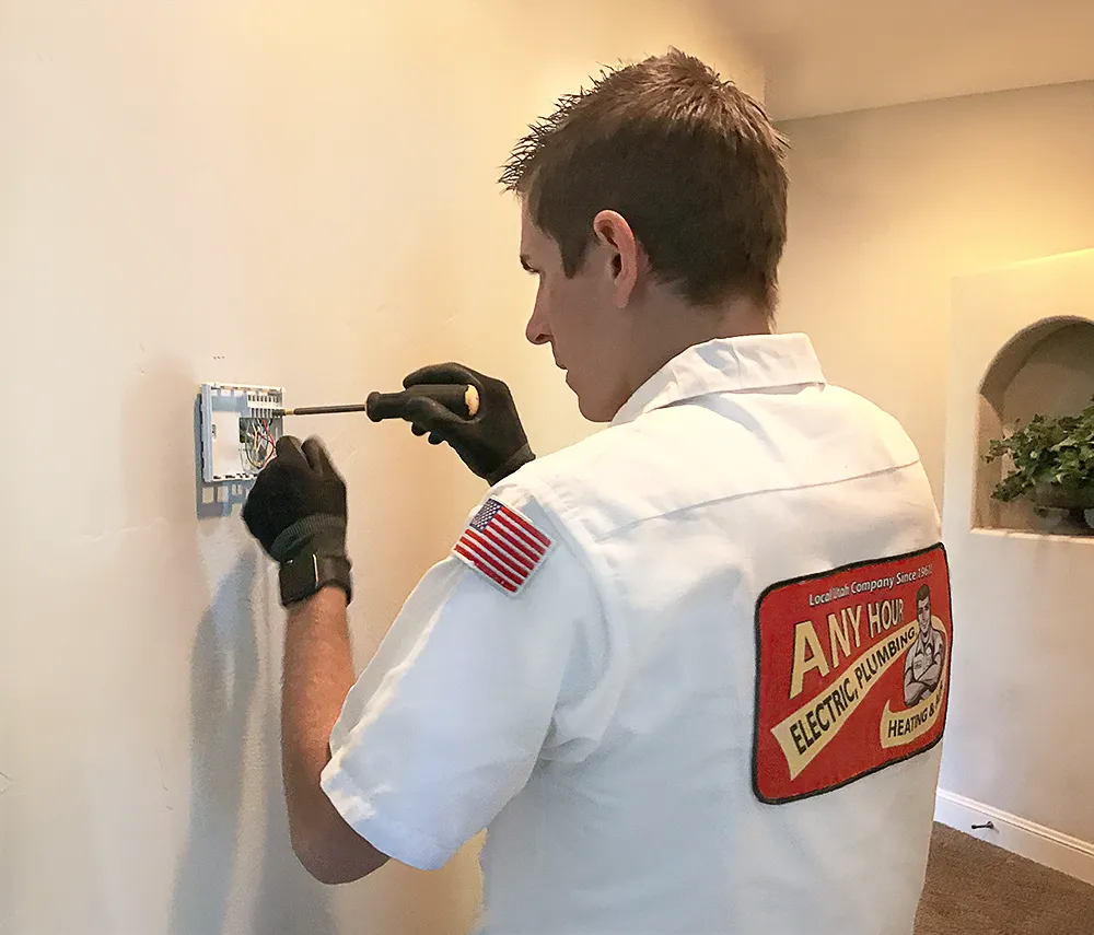 A technician in a white uniform installs a thermostat on a wall. He uses a screwdriver and wears gloves. The uniform has a patch with company details.