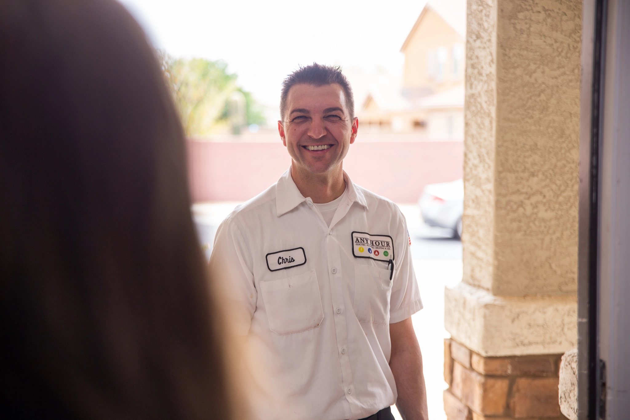 A smiling man in a white work uniform stands at a front door. Sunlit background includes a blurred view of houses and a parked car. Friendly and inviting tone.