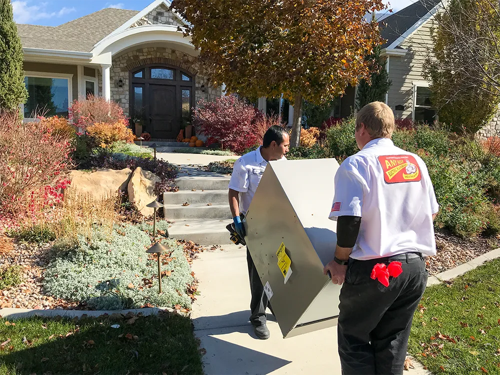 Two technicians in white uniforms carrying a furnace up a garden path. The house in the background is framed by colorful autumn foliage.