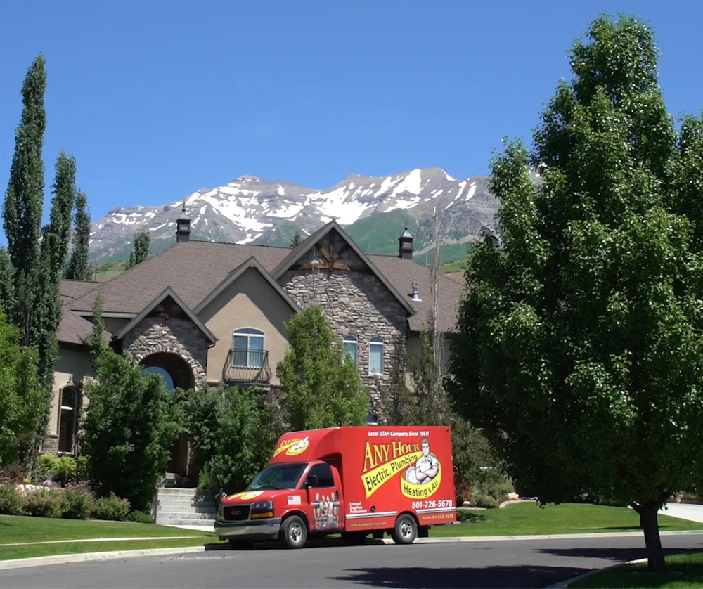 A red utility truck parked in front of a large stone and stucco house, surrounded by lush trees. Snow-capped mountains are visible under a clear blue sky.