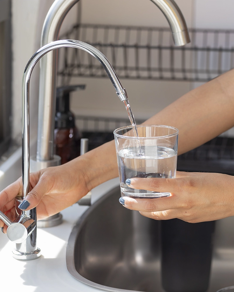 A person fills a clear glass with water from a modern kitchen faucet. The scene is bright, conveying freshness and cleanliness.