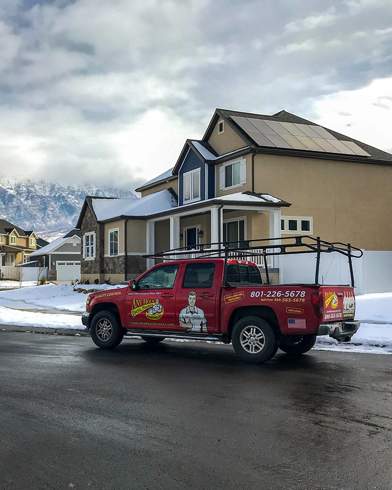 Red utility truck parked in front of a modern house with solar panels on the snowy roof, mountains and cloudy sky in the background. Winter setting.