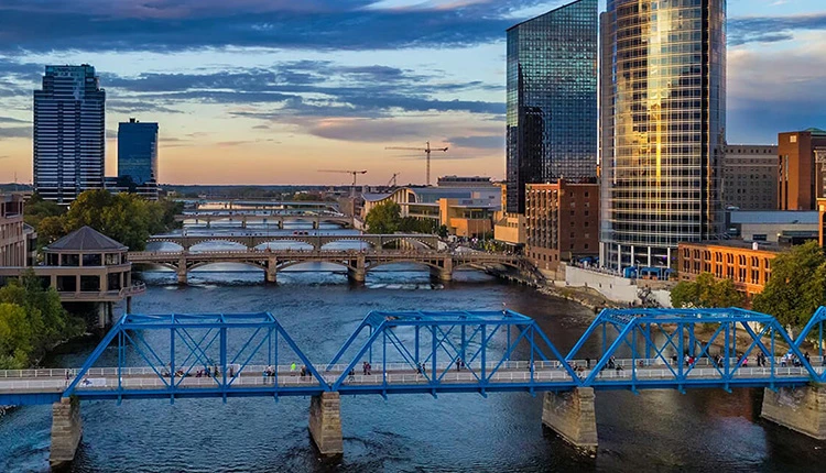 A vibrant cityscape at sunset, featuring a blue pedestrian bridge over a river, modern skyscrapers, and multiple arched bridges, evoking a serene urban atmosphere.