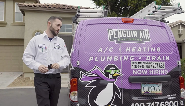 A technician in a white uniform stands beside a purple van branded Penguin Air Plumbing in front of a house. The van advertises HVAC services.