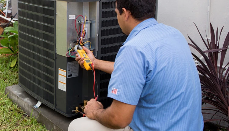 A technician in a blue shirt examines a central air conditioning unit with a multimeter. He kneels on grass, indicating an outdoor setting.