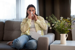 %Title% | Any Hour Services A woman sits on a sofa, holding a tissue to her nose, with a box of tissues beside her and a vase of flowers on the table in front.