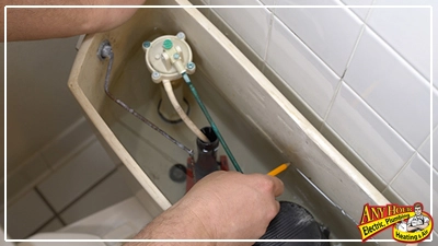 A person works to fix the toilet fill valve inside a toilet tank with the lid removed. A plumber's company logo is visible in the bottom right corner.