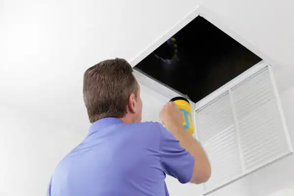 A man in a blue shirt shines a flashlight into an open attic hatch in a white ceiling, inspecting for Air Duct Repair needs in Salt Lake City.