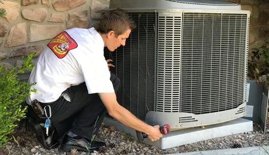 A technician in a uniform uses a power drill to service an outdoor high-efficiency air conditioner next to a building.