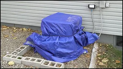 A blue tarp provides AC winter protection for an outdoor unit next to a house, secured with a cord, with gravel and cinder blocks nearby.