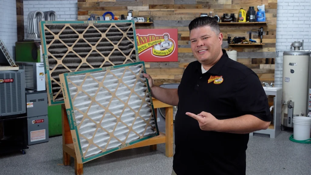 A man stands in a workshop, holding two HVAC air filters side by side, one visibly used and dirty, the other clean and new.