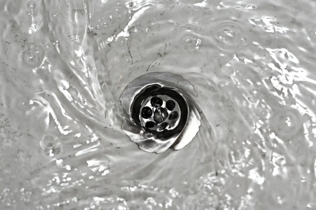 %Title% | Any Hour Services Close-up of water swirling around a metal sink drain in Salt Lake City, creating small ripples and circular motion as the water flows down—a perfect example of when a professional Drain Clearing Service might be needed.